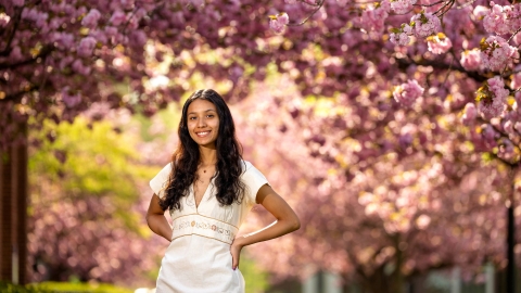 Gabby Diaz '25, in a white dress, poses with her hands on her hips in front of cherry blossom trees.