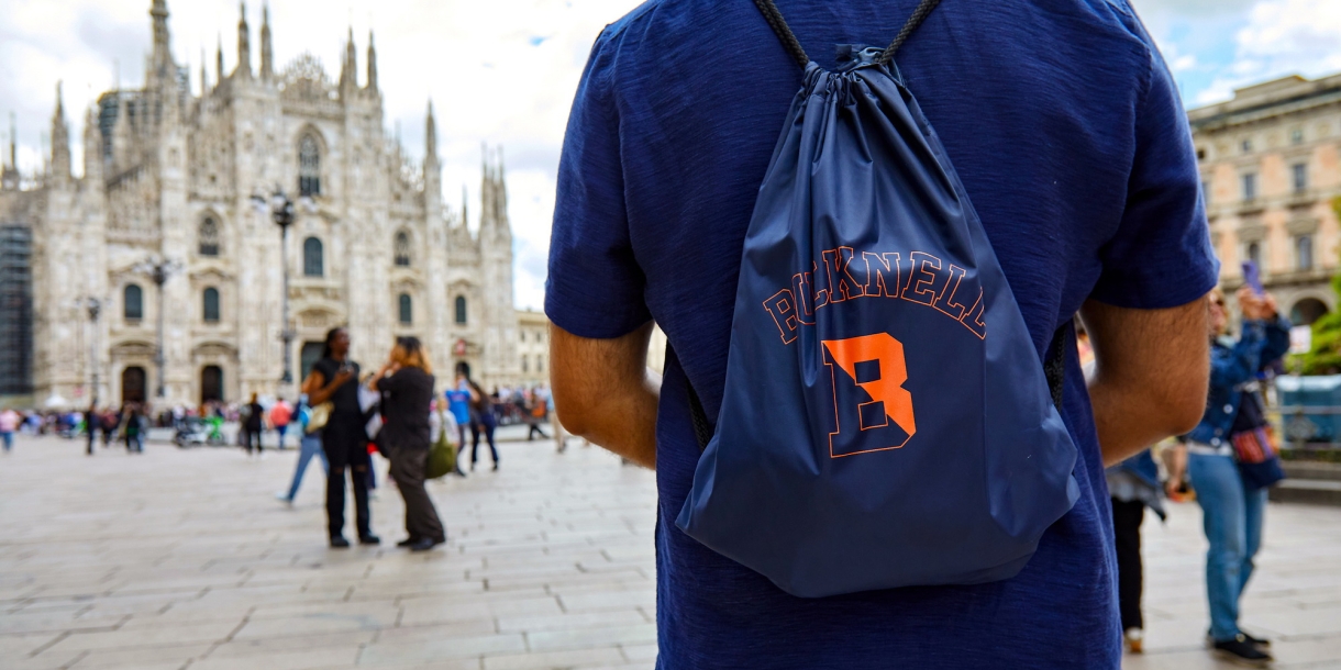 Bucknell student in a square in Milan. Student is facing away from the camera, wearing a Bucknell backpack. Cathedral in background.