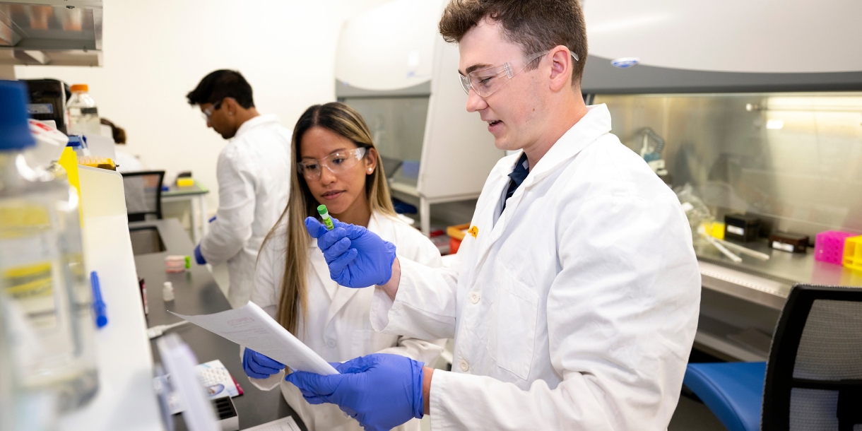 Professor Olivia Boerman and Connor Kozick '26 are wearing white lab coats and purple gloves in a lab setting.