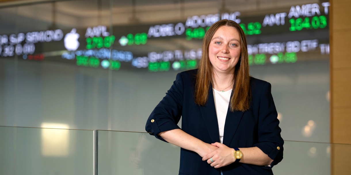Caitlin Yant '24 stands and smiles in the Moriatry Investment Center in Holmes Hall with a stock ticker behind her.
