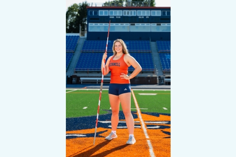 Evelyn Bliss '27 stands on the field holding a javelin dressed in her orange and blue track and field uniform.
