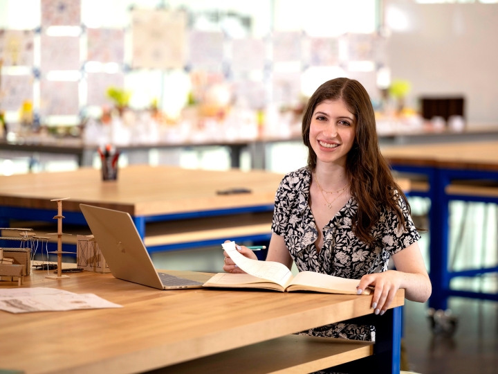 Katie Raso '28 sits in a classroom at a desk with a laptop and papers in front of her and smiles.